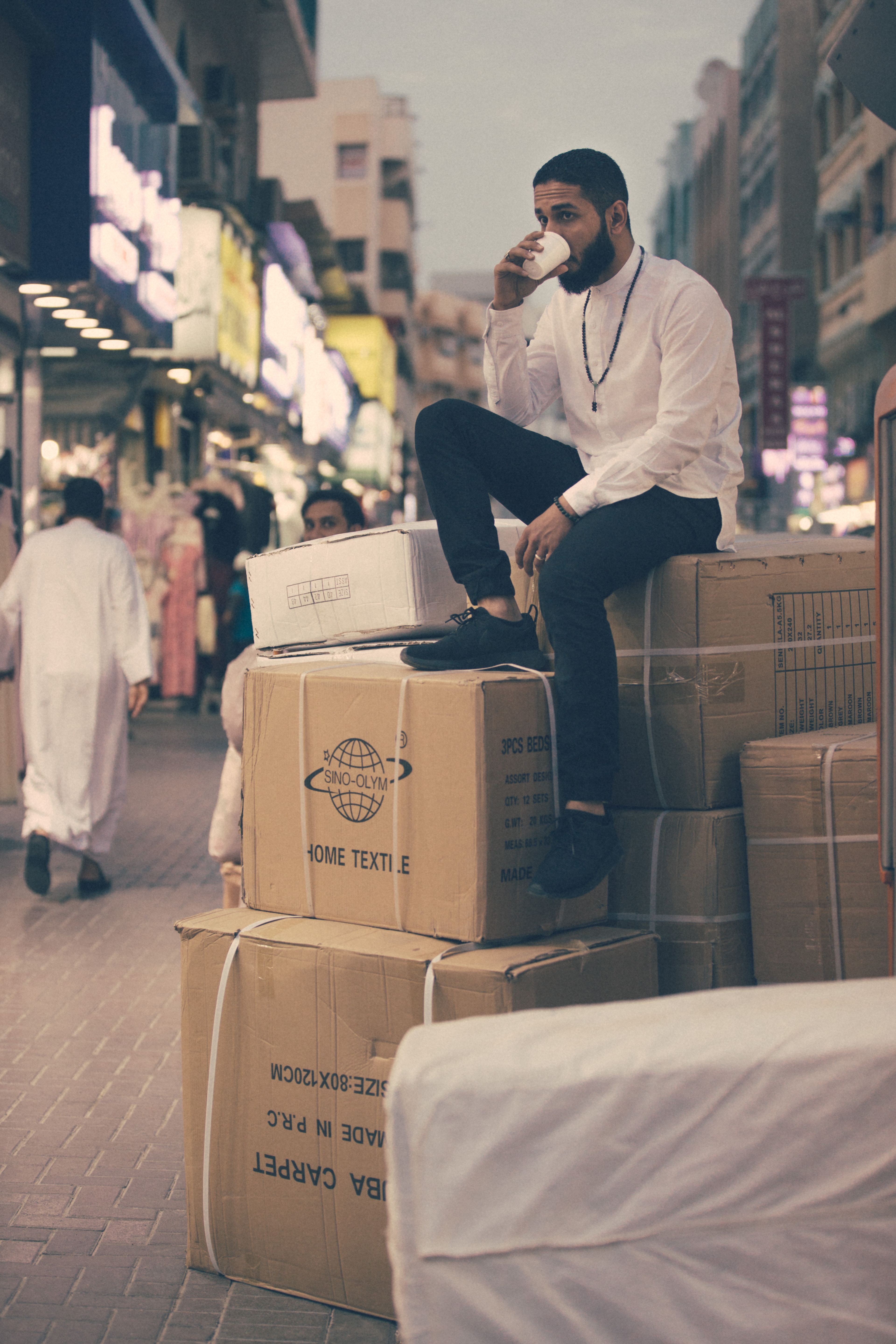 Man sipping a cup of coffee while sitting on a large stack of cardboard boxes in the street.